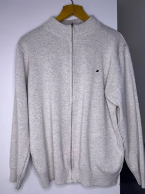 Ljusgrå ziptröja från Tommy Hilfiger - Ljusgrå stickad ziptröja från Tommy Hilfiger med liten broderad logga på bröstet. Tröjan har hög krage, hel dragkedja framtill och ribbade muddar vid ärmslut och nederkant. Perfekt för lager-på-lager och enkel att matcha.