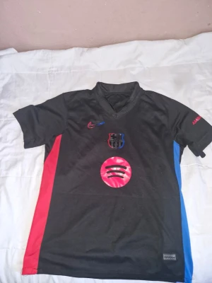 FC Barcelona tröja 24/25  - Snygg svart FC Barcelona matchtröja från Nike med klubbmärke och Spotify-logga på bröstet. Tröjan har röda och blå sidopaneler, korta ärmar och V-ringning. UNHCR-logga på ryggen och tryck på ärmarna. Perfekt för fotboll eller träning. Med ingen namn på