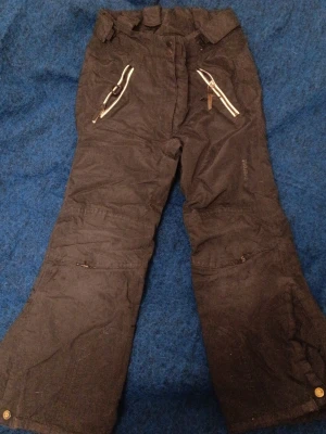 Svarta täckbyxor från Everest storlek 128 - Svarta täckbyxor med två snedställda dragkedjefickor framtill. Byxorna har breda hällor i midjan.Perfekta för kalla dagar och vinteraktiviteter. Dom har små hål i knäna. Men inget som påverkar åkningen eller lek. Storlek 128.