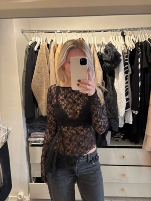 Svart spetstopp  - Säljer en svart långärmad topp i spets från Gina Tricot. Blusen är genombruten med blommigt spetsmönster och har en asymmetrisk nederkant. 