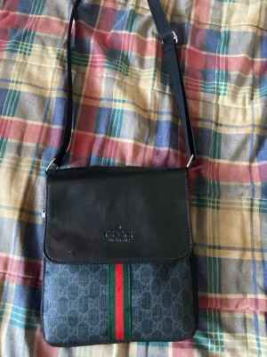 Svart axelväska från Gucci - Snygg svart axelväska från Gucci med klassiskt GG-mönster och gröna/röda ränder framtill. Väskan har justerbar axelrem, lock med präglad logga och dragkedjefack på baksidan. Perfekt för dig som gillar stilrena och ikoniska detaljer.