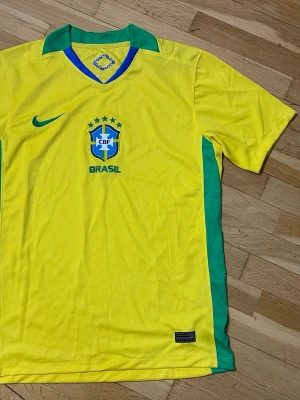 Brasilien fotbollströja  - Gul Brasilien fotbollströja från Nike med gröna detaljer och blå krage. Tröjan har CBF-logga och Nike-logga på bröstet samt Dri-FIT-material som andas. Perfekt för fotbollsträning eller att bära som supporter.