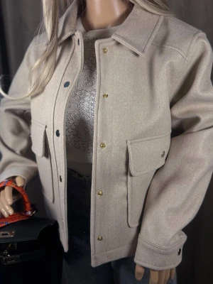 Beige overshirt med fickor M-L☑️☑️☑️ - Stilren beige overshirt med klassisk krage och stora bröstfickor med lock. Jackan har guldiga tryckknappar framtill och vid ärmslut. Tillverkad i ett mjukt, ull-liknande material som ger en lyxig känsla och passar perfekt till lager-på-lager-stil.
