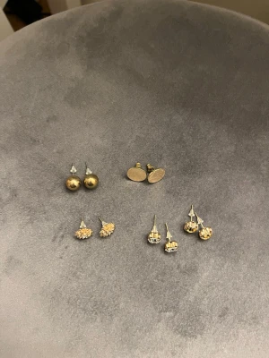 Mix av guldiga örhängen i olika former - Fem par örhängen i olika former: runda platta, klotformade, blommiga och två par med gnistrande stenar. Alla i guldton och vissa med glittriga detaljer. Perfekt för dig som vill variera din stil med klassiska och trendiga örhängen.
