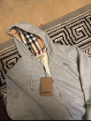 Grå zip hoodie från Burberry - Snygg grå zip hoodie från Burberry med klassiskt rutigt foder i huvan och dragsko. Hoodien har dragkedja framtill, två fickor och diskret Burberry-logga broderad på bröstet. Perfekt för en clean och stilren look. Som passar L som passar M