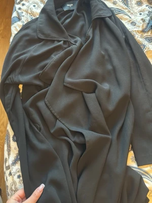 Svart lång kappa med draperad front - Stilren svart kappa från Bik Bok med snyggt draperad front och bred krage. Kappan har en avslappnad passform och är tillverkad i ett mjukt tyg som faller snyggt. Perfekt för dig som gillar en enkel men trendig look.
