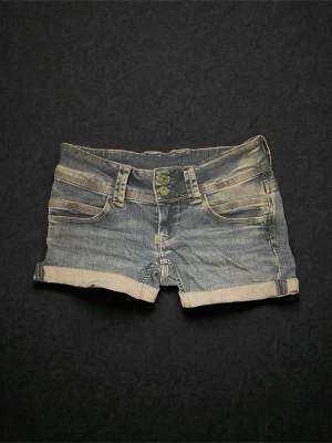 Jeans - Snygga blå jeansshorts med uppvikta benslut och dubbla knappar framtill. Klassisk femficksmodell med bälteshällor och slitna detaljer för en avslappnad look. Perfekta för varma dagar och enkel att matcha med allt.