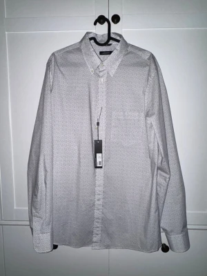 Vit skjorta med diskret mönster - Klassisk vit skjorta Lindberg med diskret småmönster, knappar framtill och bröstficka. Skjortan har lång ärm och normal passform, perfekt till både jeans och chinos. 