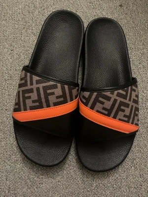 Fendi slides med logomönster och orange detalj - Säljer ett par svarta Fendi sandaler med bred rem i brunt och svart logomönster samt en snygg orange detalj. Sköna att bära med räfflad sula och klassisk öppen design. Perfekta för dig som gillar lyxiga accessoarer och streetstyle.