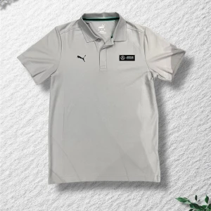 Ljusgrå pikétröja Puma Mercedes - Snygg ljusgrå pikétröja från Puma i samarbete med Mercedes AMG Petronas. Klassisk krage med knappar, korta ärmar och diskreta loggor på bröstet. Perfekt för dig som gillar sportig och stilren design.