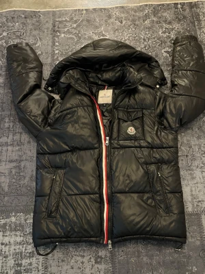 Svart dunjacka från Moncler - Snygg svart dunjacka från Moncler med huva och klassisk logga på bröstet. Jackan har dragkedja med röd, vit och blå detalj, flera fickor och quiltad design. Perfekt för kalla vinterdagar och riktigt stilren look. 