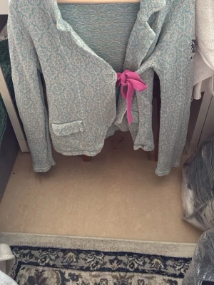 Odd Molly Ljusblå mönstrad kofta med rosett - Odd Molly  ljusblå kofta med vitt geometriskt mönster och en snygg rosa rosett i midjan. Koftan har lång ärm, en ficka framtill och mjukt material som känns skönt mot huden. Perfekt för dig som vill ha något annorlunda och stilrent i garderoben.