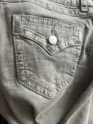 Grå jeans med broderad ficka Gina Tricot - Snygga grå jeans från Gina Tricot med unik broderad detalj på bakfickan och en dekorativ knapp. Jeansen har klassisk femficksmodell och är tillverkade i ett mjukt denimtyg. Perfekta för dig som vill ha något extra till din outfit.