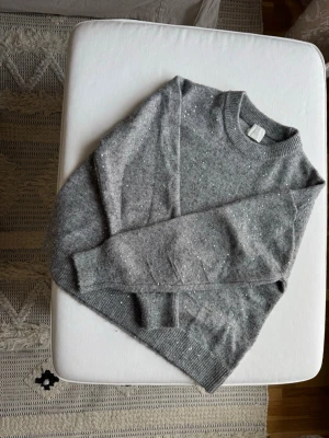 Grå stickad tröja med glitter - Mysig grå stickad tröja med diskreta glitterdetaljer. Tröjan har rund halsringning, långa ärmar och ribbade muddar vid ärmslut och nederkant. Perfekt för dig som vill ha något enkelt men ändå lite extra.
