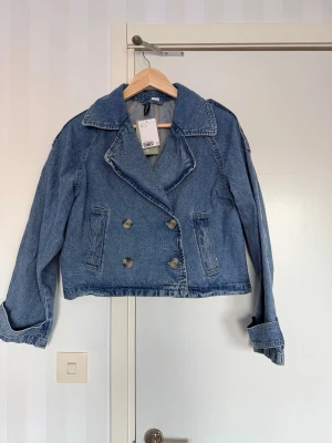 Blå cropped jeansjacka med dubbelknäppning - Snygg blå jeansjacka med bred krage och dubbelknäppning framtill. Jackan är croppad och har långa ärmar med uppvikta muddar. Perfekt för en trendig och avslappnad look.