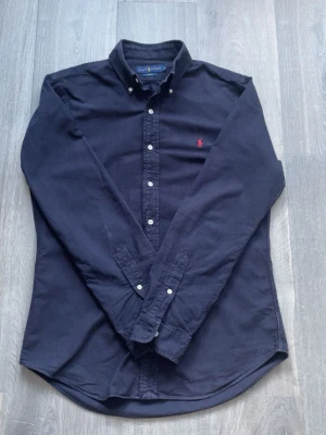 Ralph Lauren skjorta - Klassisk marinblå skjorta från Ralph Lauren. Fettfläck (sista bilden) som ej märks vid användning, annars i fint skick. Tveka inte att skriva vid funderingar✅