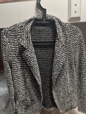 Svartvit bouclé kavaj - Stilren svartvit kavaj i bouclé-tyg med klassisk krage och lång ärm. Kavajen har en strukturerad känsla och passar perfekt till en trendig look. Inga knappar framtill och två fickor. Perfekt för dig som gillar en chic och modern stil.