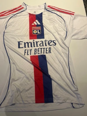 Olympique Lyonnais matchtröja Adidas - Snygg vit matchtröja från Olympique Lyonnais med röda och blå detaljer, vertikal röd/blå rand och klubbmärke på bröstet. Adidas-logga och röda ränder på axlarna. Tillverkad i lätt och ventilerande material, perfekt för fotboll eller träning.