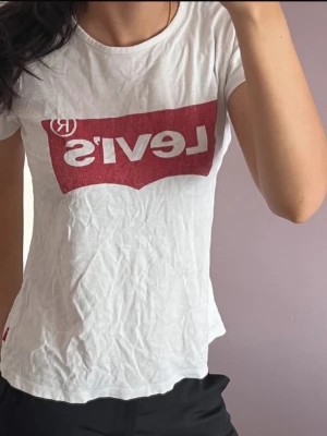 Vit Levi's t-shirt med röd logga - Vit t-shirt från Levi's med den klassiska röda loggan tryckt framtill. Modellen är kortärmad och har en rund halsringning. Tillverkad i mjuk bomull, perfekt för en avslappnad och trendig stil.