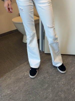 Vita bootcut jeans med slitningar - Säljer ett par vita bootcut jeans med slitna detaljer på låren och knäna. Jeansen har broderade bakfickor och en klassisk femficksmodell. Perfekta för dig som vill ha en avslappnad men trendig look.