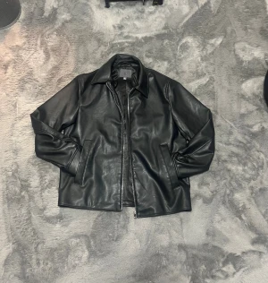 Svart skinnjacka med klassisk krage - Stilren svart skinnjacka från COS med klassisk krage och raka fickor. Jackan har dragkedja framtill och ett slätt, glansigt innerfoder. Perfekt för dig som gillar minimalistisk och tidlös stil. Det är små repor på jackan.