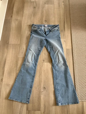 Ljusblå bootcut jeans Gina 14+ - Säljer ett par ljusblå bootcut jeans med klassisk femficksdesign och låg midja. Jeansen har snygga slitningar framtill och är tillverkade i ett mjukt denimtyg som ger en avslappnad look. Perfekta för dig som gillar retrostil och vill ha ett par jeans med lite vidare ben. Innerbenslängd: 90