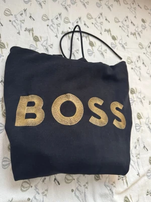 Svart hoodie från Hugo BOSS med gulddetaljer - Snygg svart hoodie från Hugo BOSS med stort guldfärgat BOSS-tryck på bröstet. Hoodien har en huva med gul insida och matchande gulddetaljer på kanten. Klassisk känguruficka framtill och ribbade muddar vid ärmar och nederkant. Perfekt för en trendig och avslappnad stil. Storlek S 