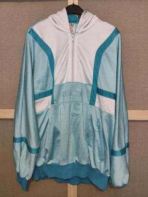 Vintage Adidas West Germany hoodie – Trefoil / 80–90-tal - Vintage Adidas hoodie från tiden då plaggen fortfarande märktes West Germany, vilket placerar den i slutet av 80-talet eller tidigt 90-tal. Det märks både i materialvalet och i konstruktionen – tyget är kraftigt, lite blankt och har den där mjuka men stadiga känslan som äldre Adidas-plagg ofta har. Färgkombinationen i ljusblått, vitt och turkos ger ett väldigt tydligt retrointryck, och den ton-i-ton-broderade adidas-loggan framtill är diskret men karaktäristisk