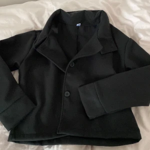 Svart kort kappa med bred krage - Stilren svart kappa med bred krage och två stora knappar framtill. Modellen är kortare och har raka ärmar. Perfekt för dig som gillar minimalistisk och klassisk stil. Materialet ser ut att vara mjukt och något tjockare, vilket ger en snygg siluett.