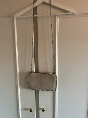 Beige kuvertväska med kedja - Snygg beige kuvertväska i mockaimitation med silverfärgad kedja som axelrem och detaljer runt kanterna. Väskan har en rektangulär form och stängs med dragkedja upptill. Perfekt för dig som gillar stilrena accessoarer med en twist.