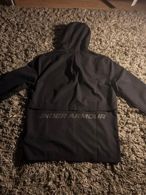 Svart vindjacka från Under Armour - Snygg svart vindjacka från Under Armour med huva och dragkedja framtill. Jackan har diskret logga på bröstet och stor logotyp på ryggen. Tillverkad i ett lätt och vindtätt material, perfekt för blåsiga dagar.