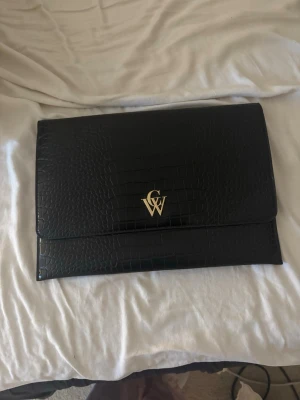 Svart kuvertväska från CW - Stilren svart kuvertväska i croco-mönstrat konstläder från CW. Väskan har guldfärgad logga på framsidan och magnetlås. Insidan är fodrad med mönstrat tyg och har en innerficka. Perfekt för skoldatorn. 36*26  