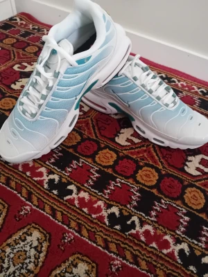 Nike Air Max Plus vita/blå sneakers - Nike Air Max Plus sneakers i vitt och ljusblått med svarta detaljer. Ovandel i mesh och syntet, klassisk vågig design och synlig Air Max-dämpning i sulan. Skorna har vita snören och svart yttersula med turkosa inslag. Perfekta för dig som gillar sportig stil.
