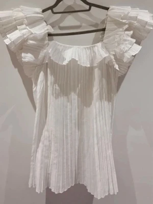 Vit plisserad klänning med volang - Tryck på köp nu💕Superfin vit klänning med plisserat tyg och stora volangärmar. Klänningen har en luftig känsla och är perfekt för varma dagar. Den har en kort längd och är ärmlös med breda axelband och volangdetaljer.