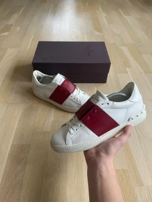 Valentino vita sneakers med röd detalj - Snygga vita sneakers från Valentino med bred röd läderdetalj över vristen och nitar på hälen. Skorna har klassisk låg profil och är tillverkade i slätt läder. Perfekta för dig som vill ha en stilren men ändå unik look.