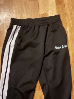 Palm Angels byxor - Svarta mjukisbyxor från Palm Angels med vita sidoränder och logga på vänster sida. Byxorna har elastisk midja och raka ben. Perfekta för en avslappnad och trendig streetwear-look.