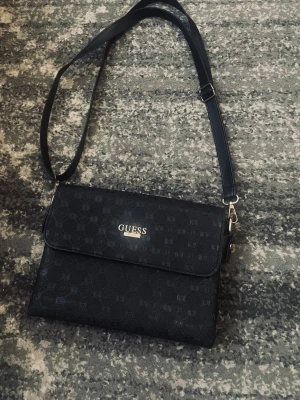 Svart axelväska från Guess - Snygg svart axelväska från Guess med justerbar axelrem och guldfärgad logga på framsidan. Väskan har ett diskret mönster med små G-detaljer och stängs med lock. Perfekt för dig som gillar stilrena accessoarer.