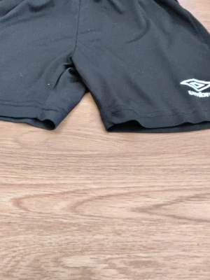 Svarta shorts från Umbro - Svarta sportiga shorts från Umbro med elastisk midja och snörning. Perfekta för träning eller fotboll. Klassisk design med Umbro-logga på ena benet. Lätta och bekväma att ha på sig.