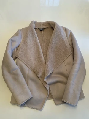 Beige teddyjacka från Gina Tricot - Supermjuk och trendig beige teddyjacka från Gina Tricot med oversized krage och öppet avslut framtill. Jackan har en mysig teddyinsida och mockaliknande utsida, perfekt för lager-på-lager-stil under kyliga dagar.