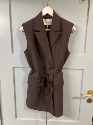 Brun väst med knyt från YAS - Stilren brun väst från YAS med dubbelknäppning, breda slag och knytband i midjan. Västen har fickor framtill och en rak, längre passform helt ny med prislappen kvar