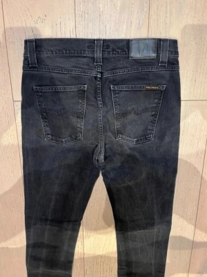 Nudie Jeans  - Säljer ett par svarta jeans från Nudie Jeans med klassisk femficksdesign och diskreta slitningar. Jeansen har en smal passform och är tillverkade i stretchigt denimtyg för extra komfort. Perfekta för en stilren och avslappnad look.