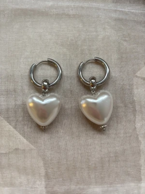 Silverörhängen med hjärtformade pärlor - Snygga örhängen i silverfärg med hjärtformade pärlor som hänger från små ringar. Perfekt accessoar för att ge din look en söt och trendig touch. Hjärtana har en pärlskimrande yta och örhängena är i metall.