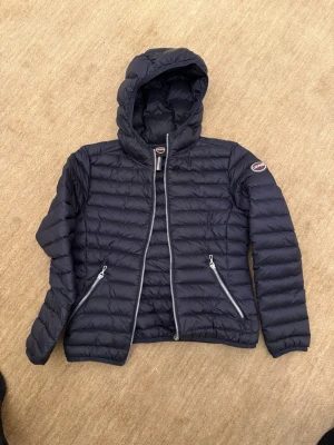 Svart dunjacka med huva från Colmar - Snygg svart dunjacka från Colmar med fast huva. Jackan har två dragkedjeförsedda fickor framtill och logotyp på ärmen. Perfekt för kalla vinterdagar och har en modern, smal passform.