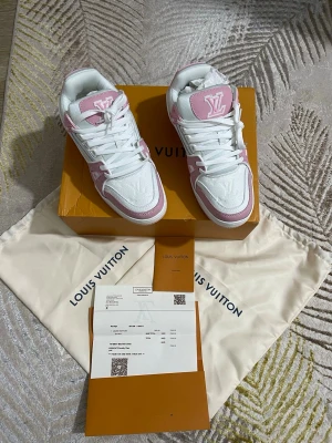  rosa och vita sneakers - Säljer ett par  sneakers i vitt och ljusrosa med klassiska LV-loggor på tungan och sidan. Skorna har en låg siluett, vita snören och detaljer i läder och textil. Perfekta för dig som vill sticka ut med exklusiva och trendiga sneakers.