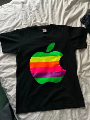 Svart Apple t-shirt med regnbågslogo - Cool svart t-shirt från Apple med stor regnbågsfärgad Apple-logga på framsidan och en mindre grön logga på ryggen. Klassisk passform och korta ärmar, perfekt för dig som gillar tech och retrostil.