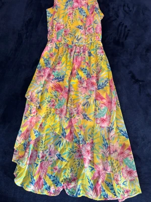 Gul blommig sommarklänning - Färgglad långklänning med blommigt mönster i rosa, grönt och blått på gul botten. Barnstorlek 152. Klänningen har ärmlös design och volangdetaljer längs kjolen. Perfekt för soliga dagar och har en luftig passform.