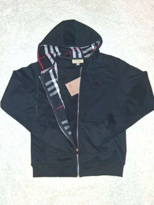 Svart hoodie från Burberry med dragkedja - Svart hoodie från Burberry med dragkedja, klassisk rutmönstrad insida på huvan och broderad logga på bröstet. Mjuk bomullskvalitet, dragsko i huvan och känguruficka framtill. Perfekt för en stilren och bekväm look.