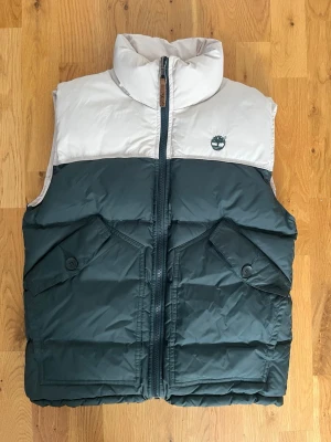 Vit och mörkgrön dunväst Timberland - Säljer en fet dunväst från Timberland i vit och mörkgrön färg. Timberland-logga broderad på bröstet. Perfekt lager på lager-plagg när det är kyligt ute.