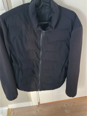 Svart pufferjacka med dragkedja - Säljer en svart cardigan med hög krage och hel dragkedja framtill. Jackan har en clean look utan synliga loggor, raka ärmar och lätt vadderad känsla. Perfekt i alla väder med sin stilrena lockbete den enkel att matcha med olika outfits.