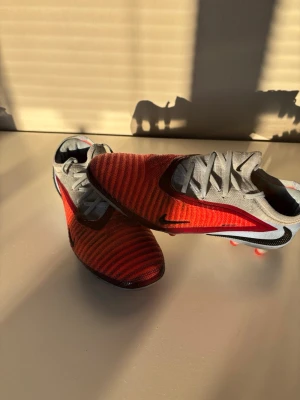 Nike Phantom fotbollsskor röd/grå - Säljer ett par Nike Phantom fotbollsskor i rött och grått med svarta detaljer. Skorna har snörning, stickad ovandel och mönstrad struktur för bättre bollkontroll. Sulan har fasta dobbar i orange för gräsplan. Perfekta för dig som vill sticka ut på planen.